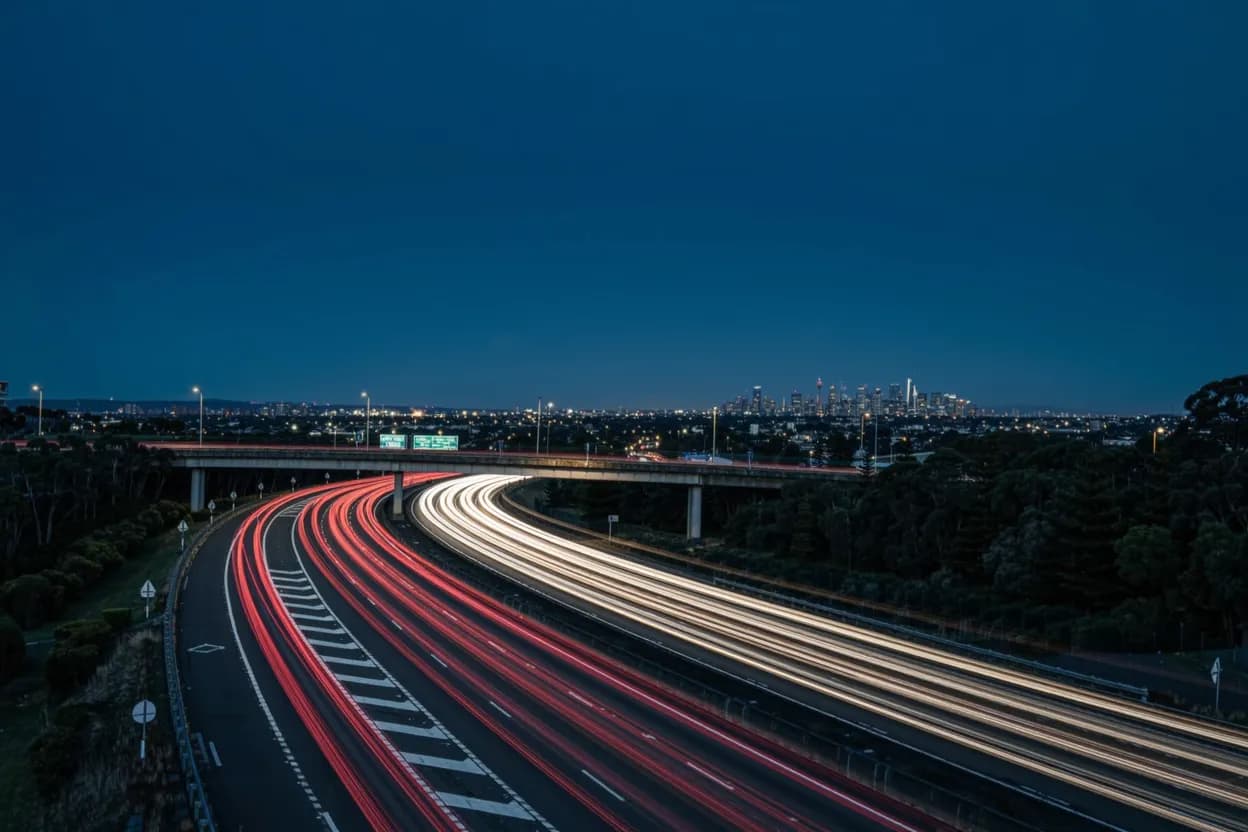 Light trails on a wide multi-lane Melbourne freeway at dusk, the city skyline glowing on the horizon